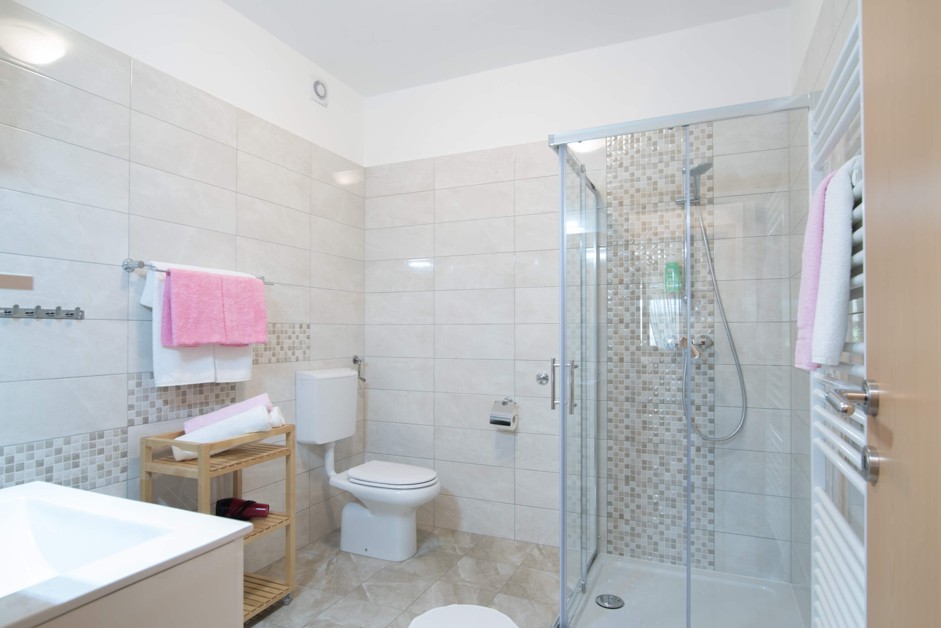 Big tiled spacious bathroom with a sink, towels, toilet, shower and radiator. Colorful towels on the hangers, radiator and towel shelf with hair dryer. Additional hangers on the wall. Toilet paper near the toilet. Shampoo in a glass shower cabin.