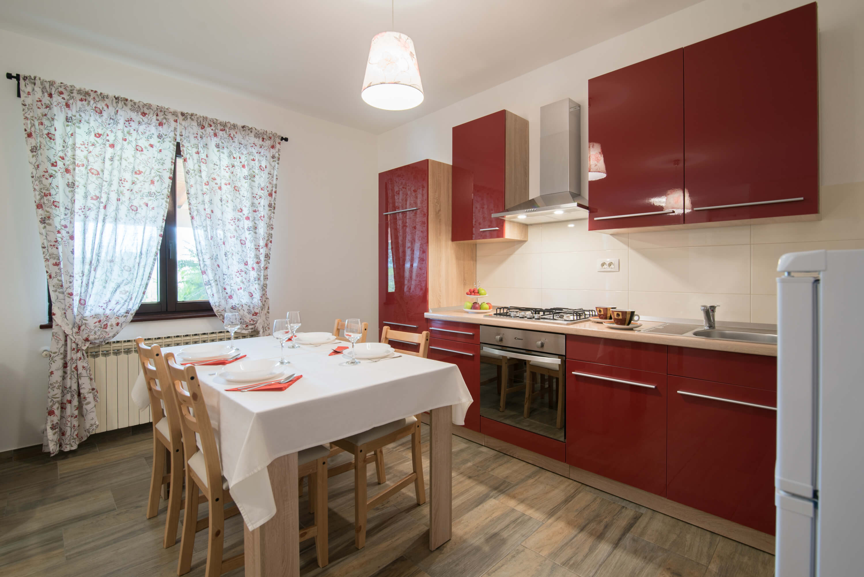 Red kitchen and bright dining room with big window. Lined dining table with tablecloth and four chairs. Big red kitchen with a big refrigerator and every possible appliance including stove, sink, and cupboards.