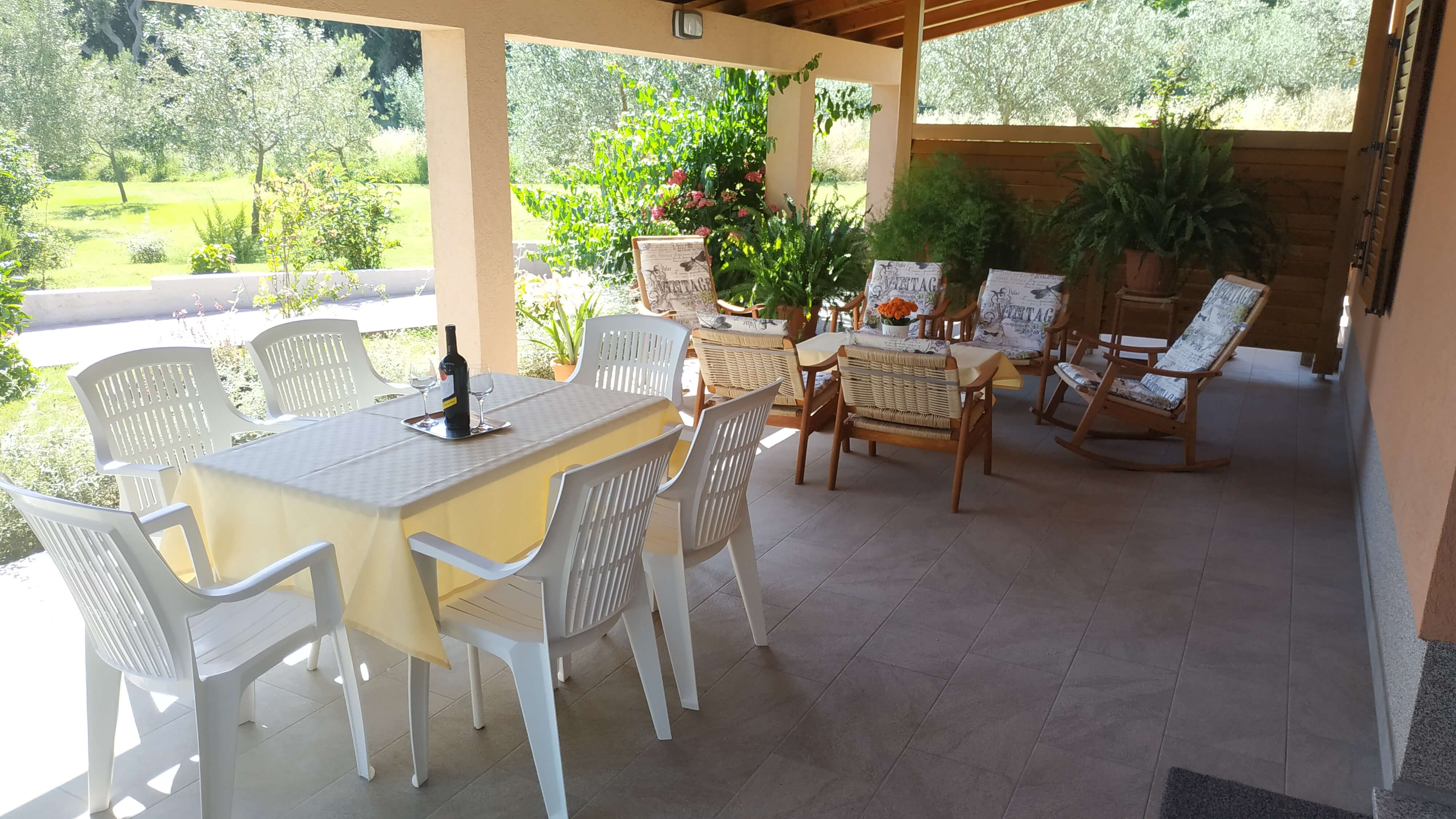 Chairs and tables on a large terrace. Dining table with plastic chairs, tablecloth and wine. Coffee table with tablecloth on it. Armchairs next to the coffee table. Big window on the wall. Wooden barrier for privacy and plants. View into the garden.